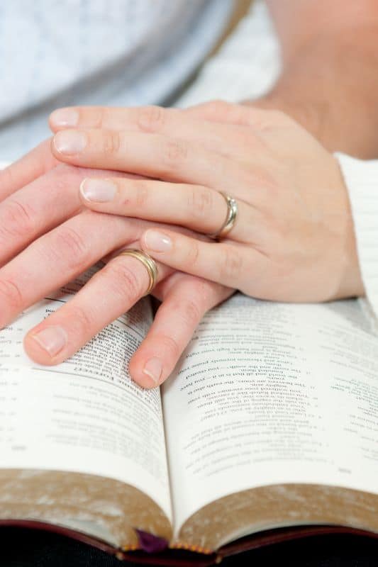 woman's hand over man's hand on Bible for war room prayers for your marriage
