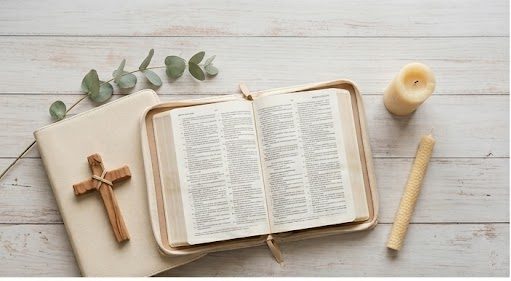An open Bible on a light wood surface next to a wooden cross, eucalyptus, and a candle, illustrating a peaceful study on understanding spiritual warfare.
